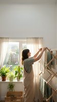 Woman with dark hair, Caucasian ethnicity, wearing casual beige clothing, hanging freshly laundered textiles on wooden drying rack indoors near bright window with sheer curtains and potted plants.
