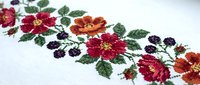 Traditional cross stitch embroidery with colorful floral pattern featuring red, orange and purple flowers with green leaves on white fabric.