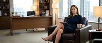 Confident Caucasian businesswoman in navy dress sitting in modern office armchair with tablet and coffee cup, city view background.