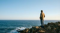 Man standing on rocky coastal cliff overlooking calm ocean waters at sunset, contemplative travel and adventure lifestyle scene for editorial and wellness content.