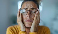 Young woman in glasses and yellow sweater experiencing headache or migraine, holding head with closed eyes showing pain and discomfort in soft lighting.