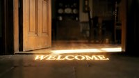 Warm golden light shining through an open wooden door onto a stone floor with a glowing welcome sign, inviting home entrance scene.
