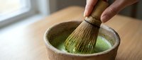 Person whisking matcha powder in ceramic bowl with bamboo whisk, preparing traditional green tea beverage for culinary and wellness purposes.