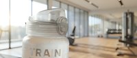 Young athletic woman with South Asian ethnicity, wearing fitness attire, holding water bottle labeled "TRAIN" in modern gym setting with equipment and windows. Hydration and workout motivation concept.
