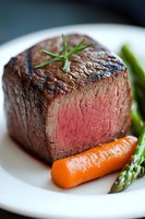 Juicy medium-rare filet mignon steak with grilled asparagus and glazed carrot on white plate, garnished with fresh rosemary. Close-up food photography.
