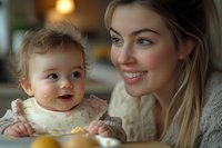 Young Caucasian mother with blonde hair smiling while feeding messy baby in high chair. Warm lighting creates cozy atmosphere during mealtime bonding moment.