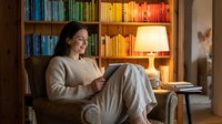 Young Caucasian woman relaxing in cozy armchair with tablet, colorful bookshelf and warm lamp light creating inviting home library atmosphere.