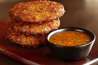 Crispy breaded potato cakes stacked on wooden plate with spicy orange dipping sauce in black ceramic bowl, close up food photography on brown background.