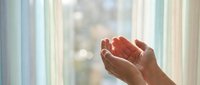 Open cupped hands reaching upward in prayer or meditation gesture against soft window light background for spiritual wellness concepts.