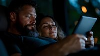 Mature bearded man and young woman looking at digital tablet screen in dark room with soft ambient lighting. Cozy evening atmosphere.