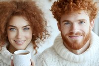 Young mixed-race woman and Caucasian man with curly red hair smiling together in cozy white sweaters, sharing warm moment with coffee cup at home.