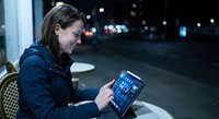 Young Caucasian businesswoman using tablet computer at night cafe with city lights background for remote work and digital lifestyle concepts.