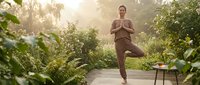 Young woman practicing yoga tree pose on wooden deck surrounded by lush tropical plants during golden hour sunrise creating peaceful wellness atmosphere.