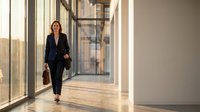 Professional businesswoman in black suit walking through modern office corridor with natural lighting and glass walls for corporate presentations.