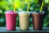 Three colorful fruit smoothies in plastic cups with yellow straws on wooden surface against blurred green background for healthy lifestyle content.