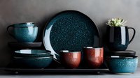 Modern ceramic dinnerware set in speckled teal blue and terracotta red colors arranged on dark background, featuring plates, bowls, cups and succulent planter.