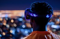 Young African American man wearing futuristic LED glasses gazes at city lights bokeh backdrop during night, orange shirt, curly hair silhouette creates modern urban atmosphere.