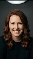 Professional portrait of smiling Caucasian businesswoman with brown wavy hair wearing dark sweater against gray background for corporate headshots.