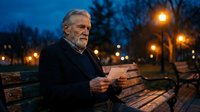 Senior Caucasian man with gray beard reading newspaper on park bench at evening twilight with warm street lights creating atmospheric urban scene.
