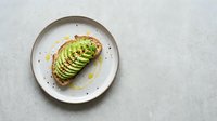 Sliced avocado on toasted bread with olive oil and seasoning on a ceramic plate, overhead view for food styling and culinary content.