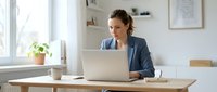 Young Caucasian businesswoman in blue blazer working on laptop at home office desk, focused on remote work tasks.
