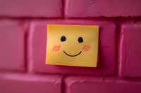 Yellow sticky note with hand-drawn happy face and rosy cheeks on pink painted brick wall background, representing positivity and cheerful messages.
