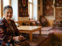 Senior woman wearing traditional Central Asian clothing and headscarf sits in ornate interior with patterned rugs and wooden furniture, representing cultural heritage and traditional lifestyle.