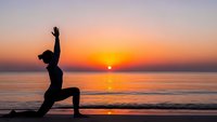 Woman practicing yoga warrior pose silhouette on beach at sunrise with golden sky and ocean waves for wellness meditation and fitness lifestyle.