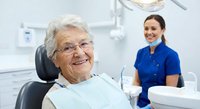 Smiling elderly Caucasian woman patient in dental chair with friendly female dentist in blue scrubs at modern clinic for oral health checkup and care.