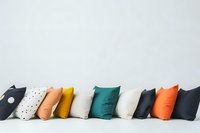 Colorful decorative throw pillows arranged in row against white wall, featuring modern geometric patterns and solid colors in teal, orange, yellow and black tones.