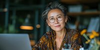 Confident mature Asian woman with gray hair and glasses working on laptop in cozy cafe environment with warm lighting.