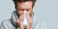 Man with gray scarf blowing nose into tissue, showing symptoms of cold or flu against light background. Concept for seasonal illness, allergies, and healthcare.