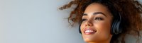 Young African American woman with curly hair and bright smile wearing wireless headphones enjoying music against light background, panoramic crop.
