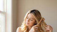 Young blonde woman gently combing her long hair with wooden comb in soft natural light by window, peaceful morning self-care routine and healthy haircare habits.