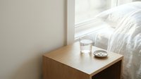 Minimalist wooden bedside table with glass of water and small bowl in bright bedroom interior with white bedding and natural light from window.
