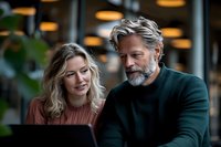 Mature businessman with gray beard and young blonde female colleague reviewing presentation on laptop in modern office space with warm lighting.