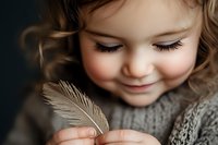 Young blonde girl in knit sweater gently holding delicate feather with closed eyes and peaceful smile. Childhood innocence and wonder concept.