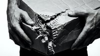 Close-up of male hands holding torn black fabric with frayed edges against white background, showing texture and damage detail.