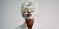 Woman with facial mask wearing cucumber slices on eyes and white towel on head during spa treatment, relaxing in bathrobe against neutral background.