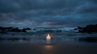 Solitary candle glowing on wet beach sand at twilight with dramatic stormy clouds and ocean waves creating peaceful meditation atmosphere.