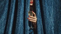 Woman with red manicured nails holding champagne glass peeking through blue velvet curtains for celebration and luxury lifestyle concepts.