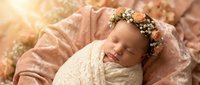 Peaceful newborn baby girl sleeping wrapped in cream knit blanket wearing delicate floral crown headband in soft warm golden sunlight for portrait photography.
