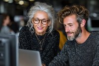 Mature woman with gray hair and glasses collaborating with bearded young man at laptop, both smiling while working in modern office environment.