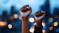 Raised fists silhouetted against blurred city lights and skyline at dusk, symbolizing protest, unity, resistance and social movement in urban setting.