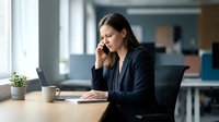 Professional businesswoman talking on phone while working on laptop in modern office environment for corporate communication and productivity concepts.