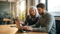 Two professionals collaborating at laptop in bright modern office with plants and windows, suitable for business, teamwork, and corporate communications.