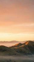 Misty rolling hills bathed in warm golden sunrise light, with layered mountain ridges fading into atmospheric haze. Serene landscape for travel, nature, and wellness projects.