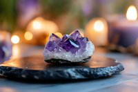 Raw amethyst crystal cluster on black stone plate with glowing candles creating magical atmosphere, purple healing gemstone in spiritual setting.