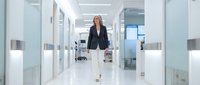Confident businesswoman with blonde hair, Caucasian ethnicity, wearing dark blazer and white pants, walking through modern office corridor with glass partitions and bright lighting.