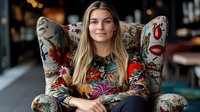 Young blonde woman in floral dress sitting in vintage patterned wingback chair, with soft bokeh background creating cozy atmosphere. Professional lifestyle portrait.